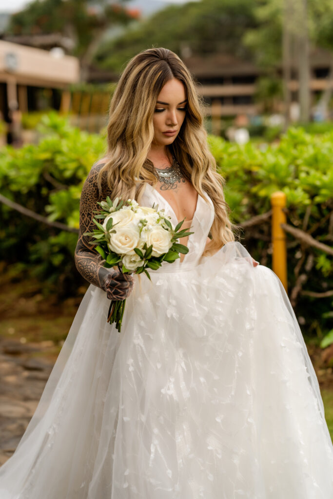 Bride in lace wedding gown with white bouquet walks path at Maui wedding venue.