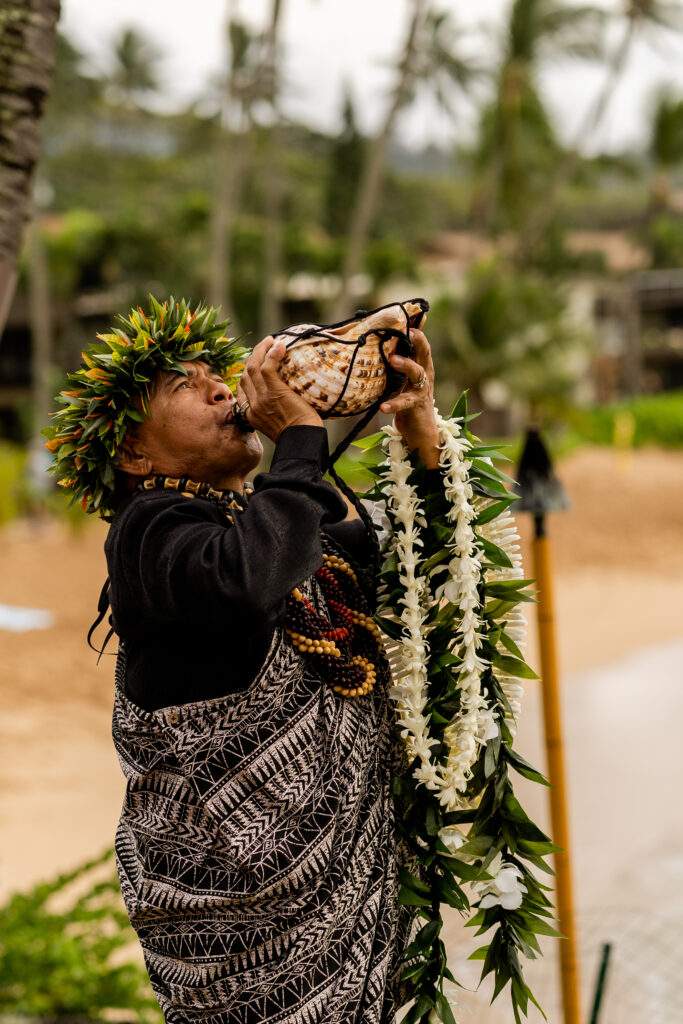 Hawaiian officiant with lei blows conch shell at Maui elopement.