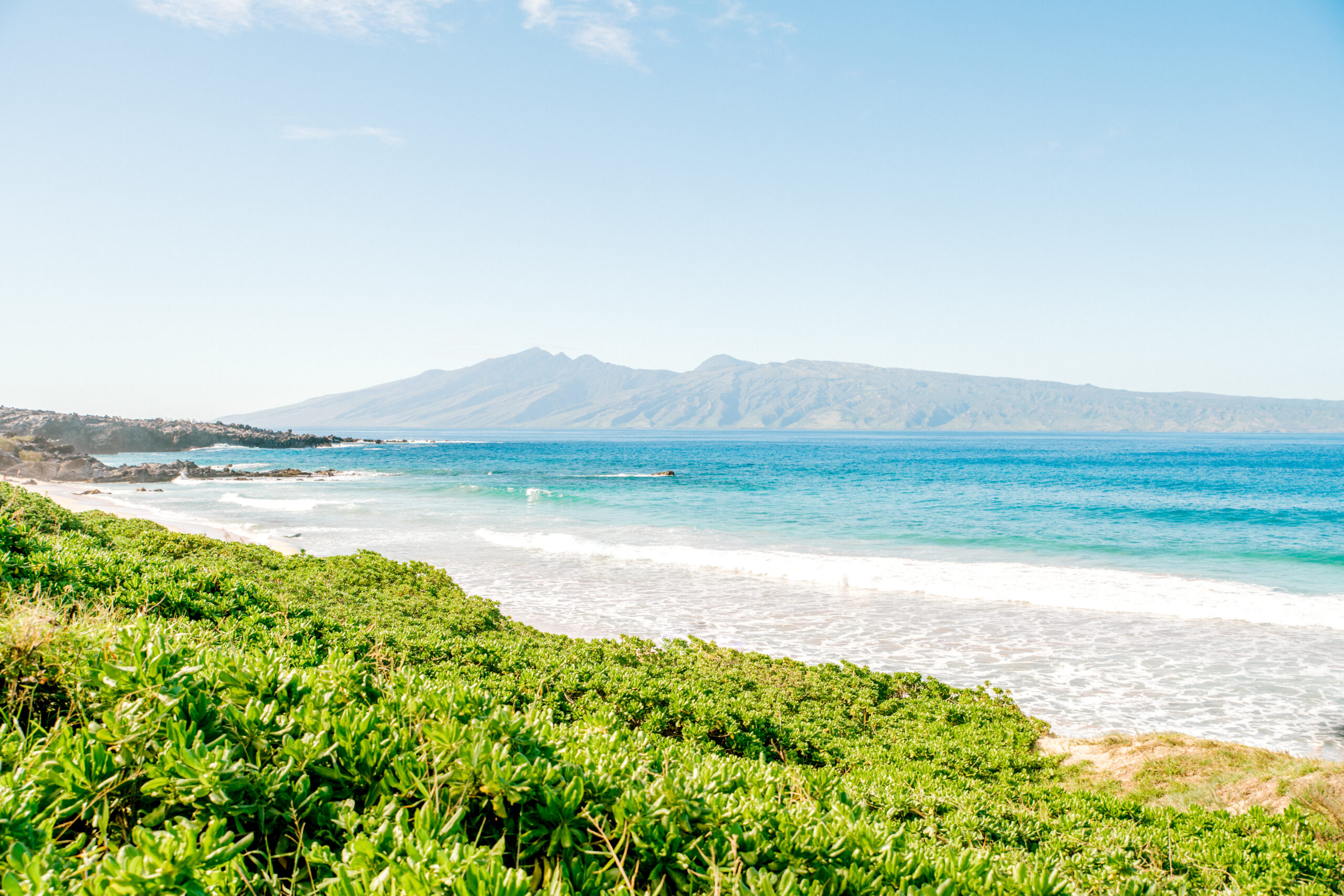 Scenic Maui beach with green shoreline, turquoise water, and distant mountains under a clear sky.
