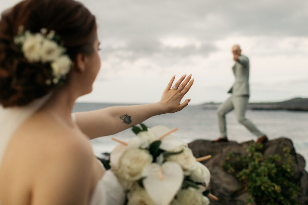 Wedding ring on the bride’s hand with a Maui beach scene and her groom in the background