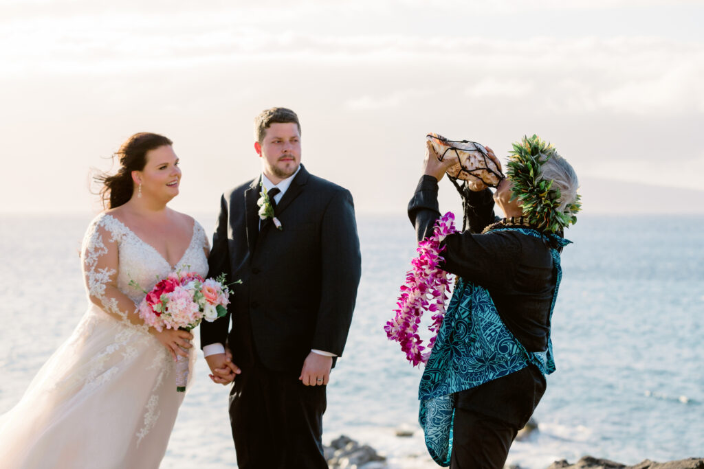 Maui cliffside wedding ceremony with Hawaiian officiant blowing conch shell as bride and groom hold hands by the ocean.