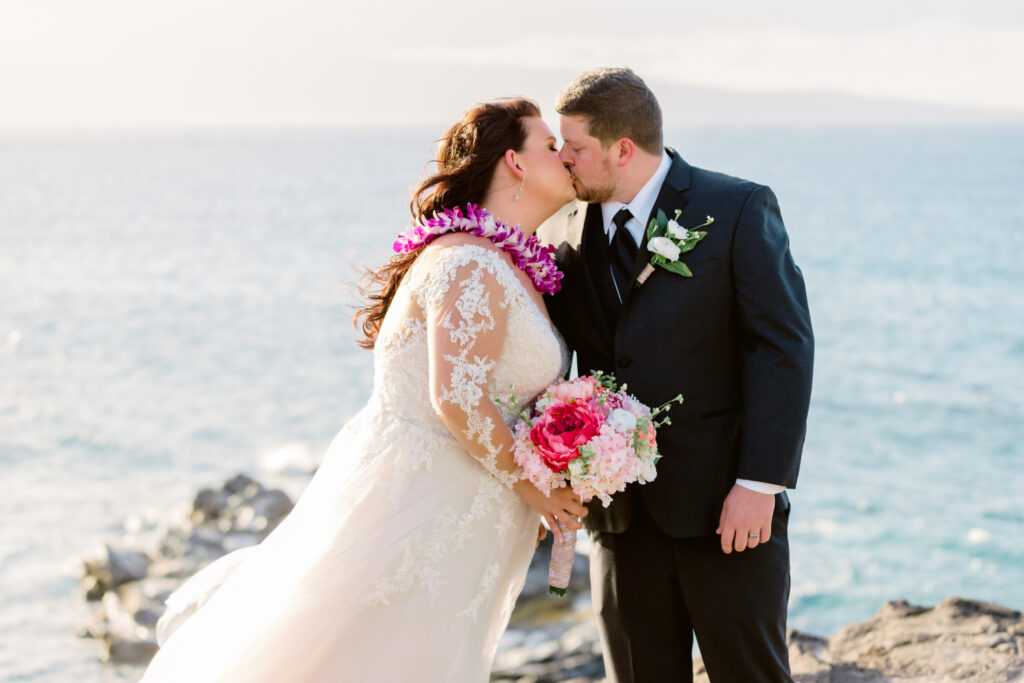 Bride and groom kissing on a rocky oceanfront cliff on Maui, bride in lace-sleeve gown with pink bouquet and orchid lei.