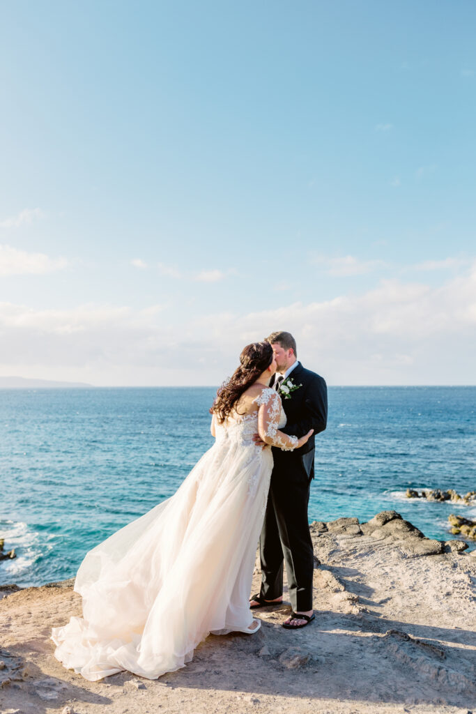 Bride and groom embracing on a sunlit cliff on Maui with waves and blue ocean in the background.