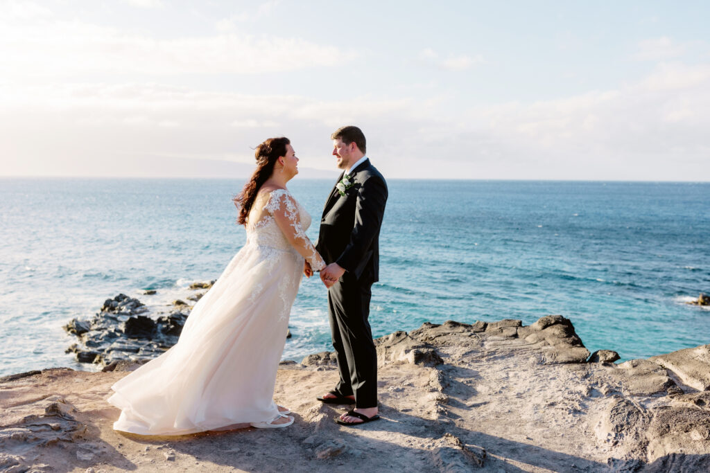 Bride and groom facing each other and holding hands on a Maui cliff, wearing purple orchid leis during their oceanfront ceremony.