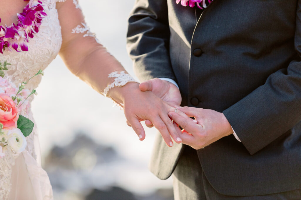 Groom placing wedding ring on bride’s finger during intimate oceanfront ceremony on a rocky cliff on Maui.