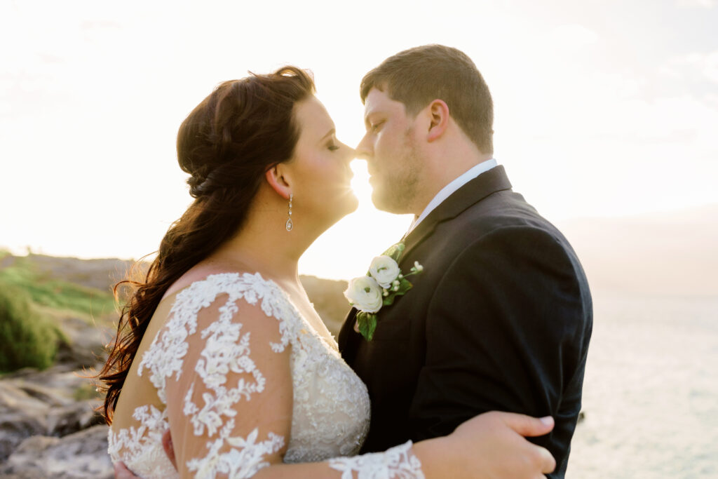 Bride and groom standing hand in hand on a rocky cliff on Maui, waves and blue ocean in the background.