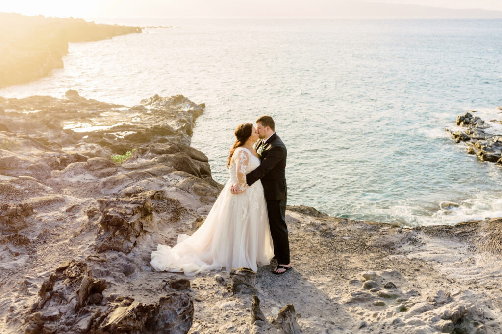 Bride and groom kissing on a rocky shoreline on Maui at sunset with golden light over the ocean.