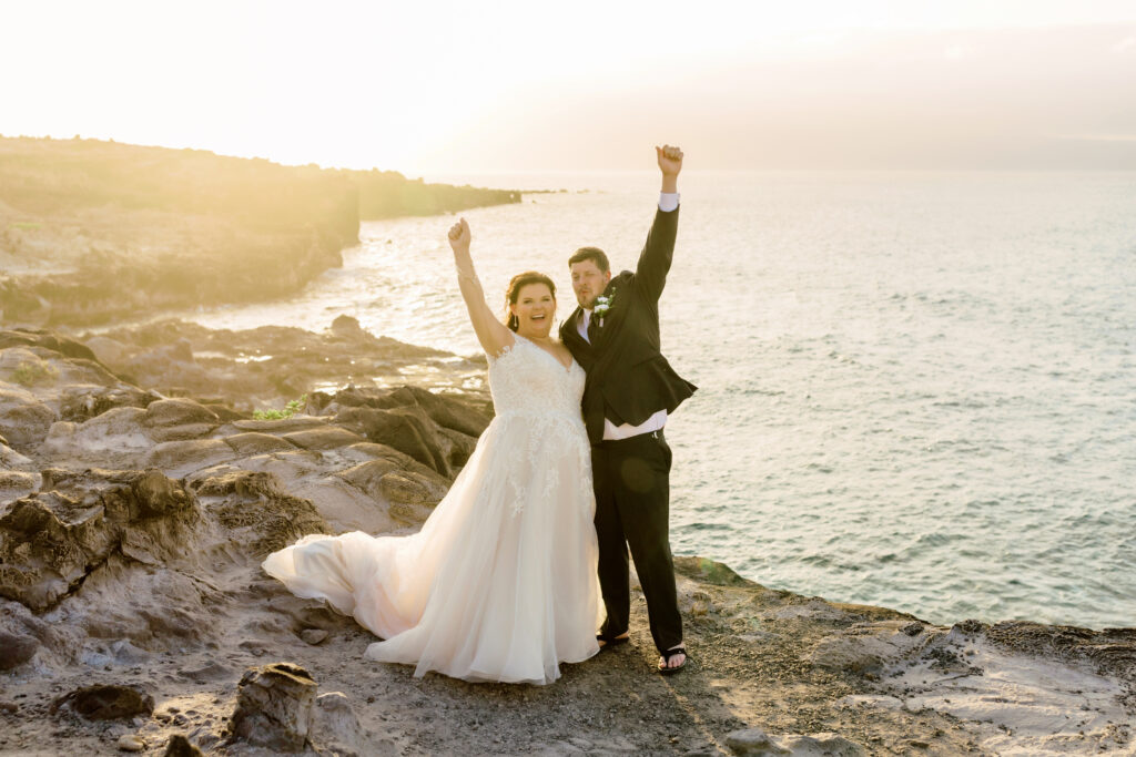 Newlywed couple on a rocky cliff on Maui at sunset, holding hands and raising their arms in celebration by the ocean.