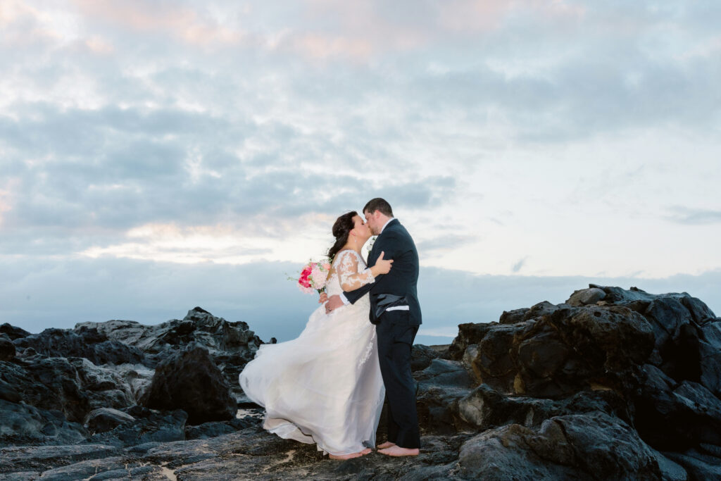 Bride and groom kissing barefoot on dark lava rocks on Maui at dusk, wedding dress flowing in the ocean breeze.