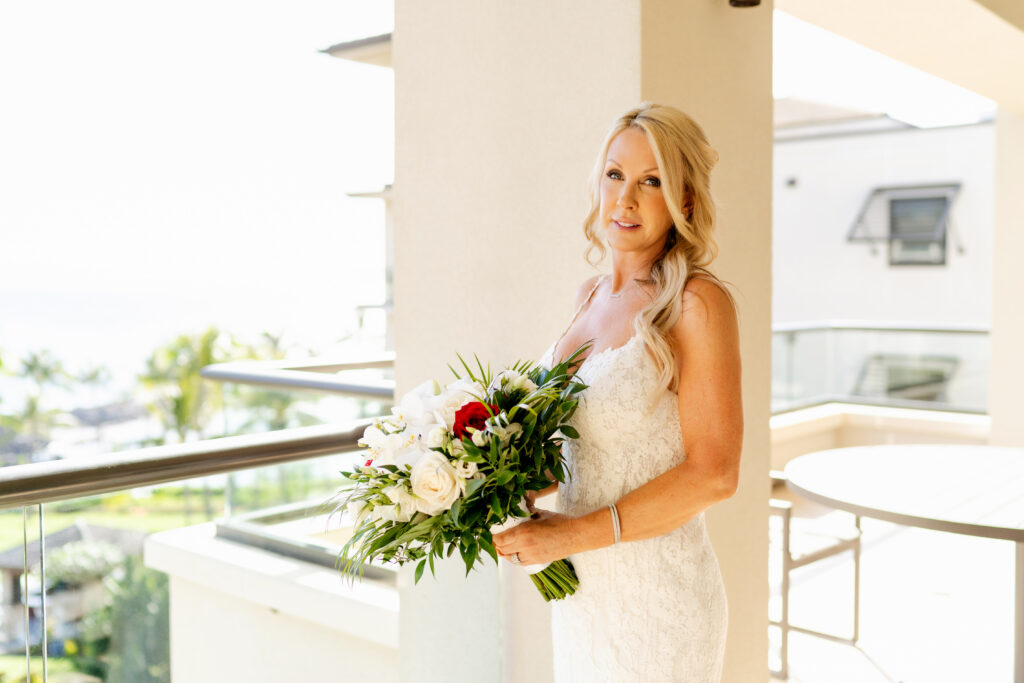 Bride in a fitted lace wedding gown holding a tropical bouquet on a bright balcony overlooking a Maui resort and ocean.

