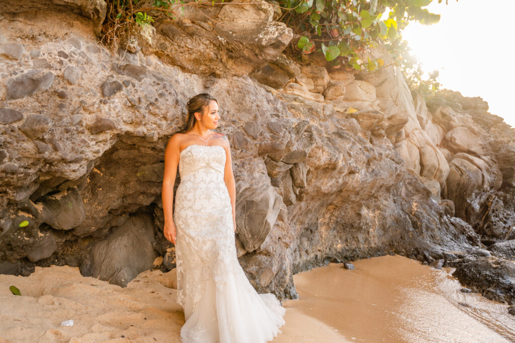 Bride in a flowing sleeveless gown standing on a cliffside overlooking the ocean at sunset, holding a bouquet and wearing a flower crown.
