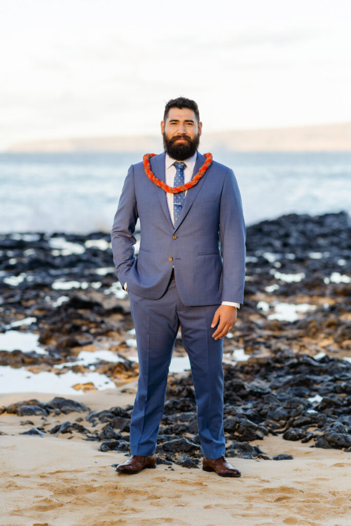 Groom in a bright blue suit and white shirt with a green maile style lei on a Maui shoreline.