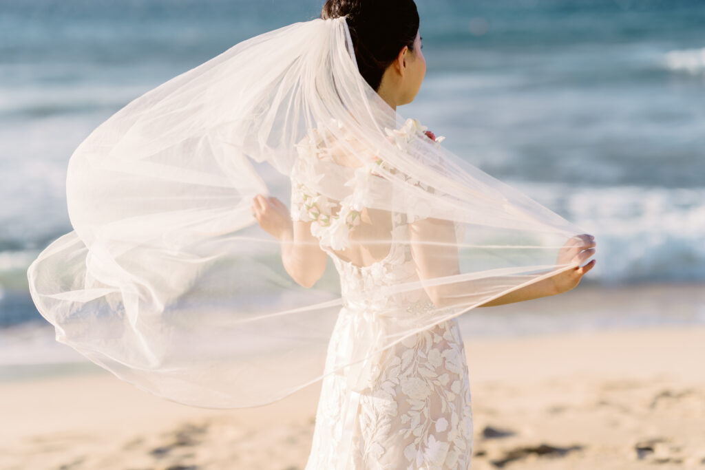Bride on a sandy Maui beach holding out her long veil as it flows in the breeze with the ocean in the background.

