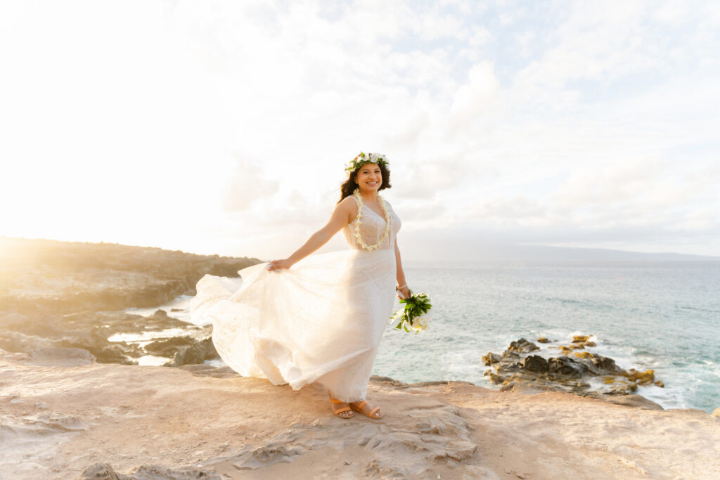 Bride in a lace wedding gown standing against a rocky cliff on a Maui beach at sunset.