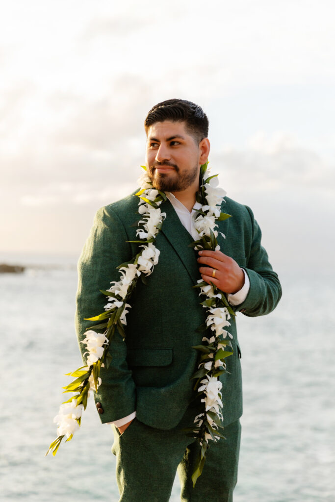 Groom in a green suit with a white orchid lei standing by the ocean at golden hour.

