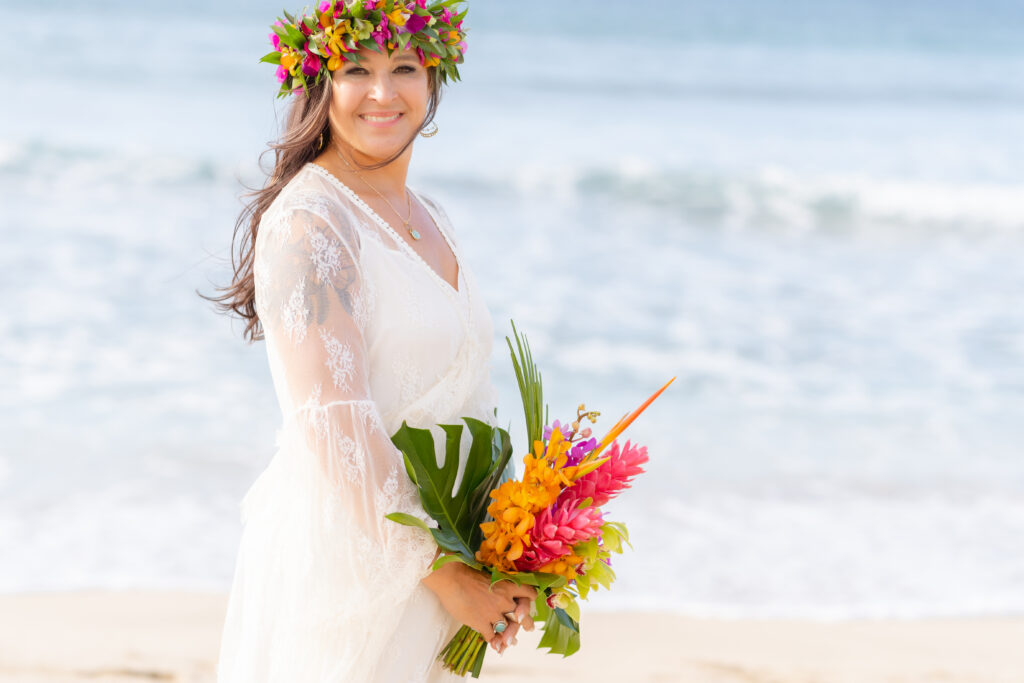 Bride in a romantic tulle wedding dress holding a bouquet of tropical flowers on a Maui beach.


