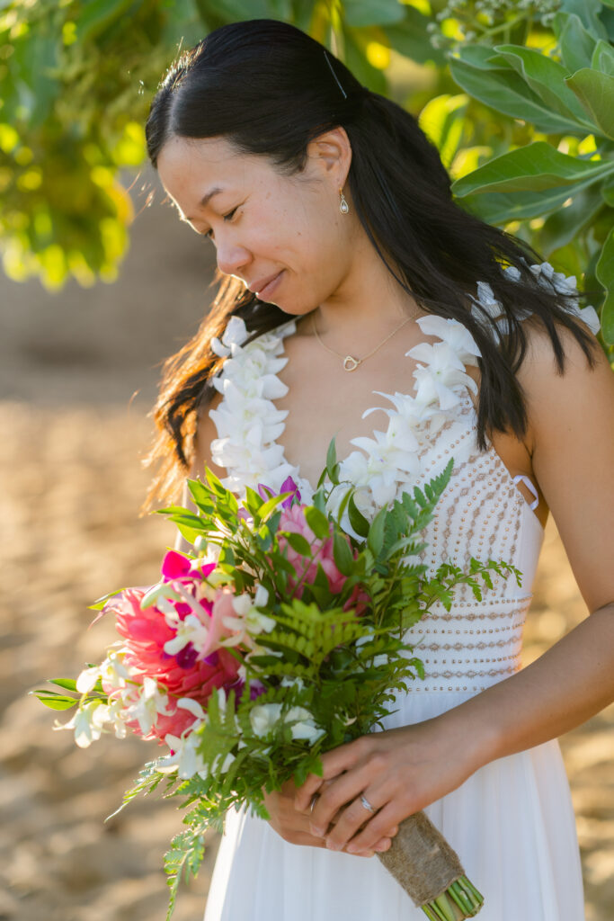 Bride in a white sleeveless dress wearing a white orchid lei and holding a bright pink and green tropical bouquet on a sandy Maui beach near greenery.

