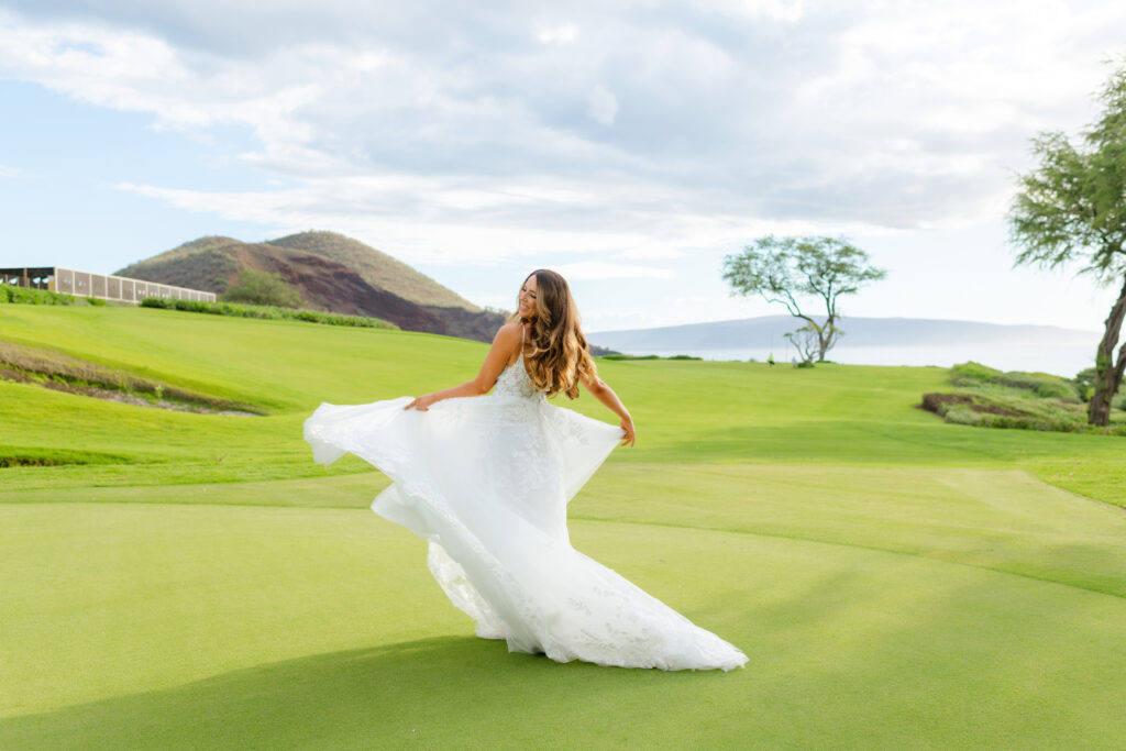 Bride in a flowing lace gown twirling on a green golf course with ocean and hills in the distance at a Maui resort.




