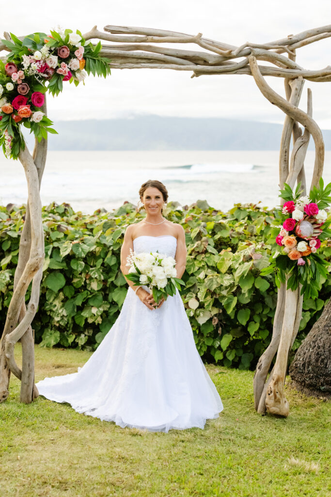 Bride in a strapless ballgown holding a white bouquet, standing under a driftwood arch decorated with colorful tropical flowers at an oceanfront Maui ceremony site.

