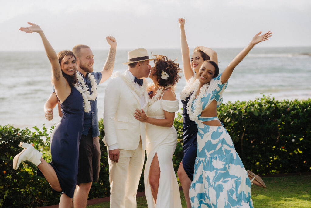 Bride and groom kissing at an oceanfront Maui venue, surrounded by cheering guests in blue outfits and white leis.