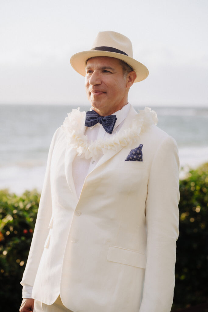 Groom wearing a white dress shirt, navy tie, and white lei on a sunny Maui beach.