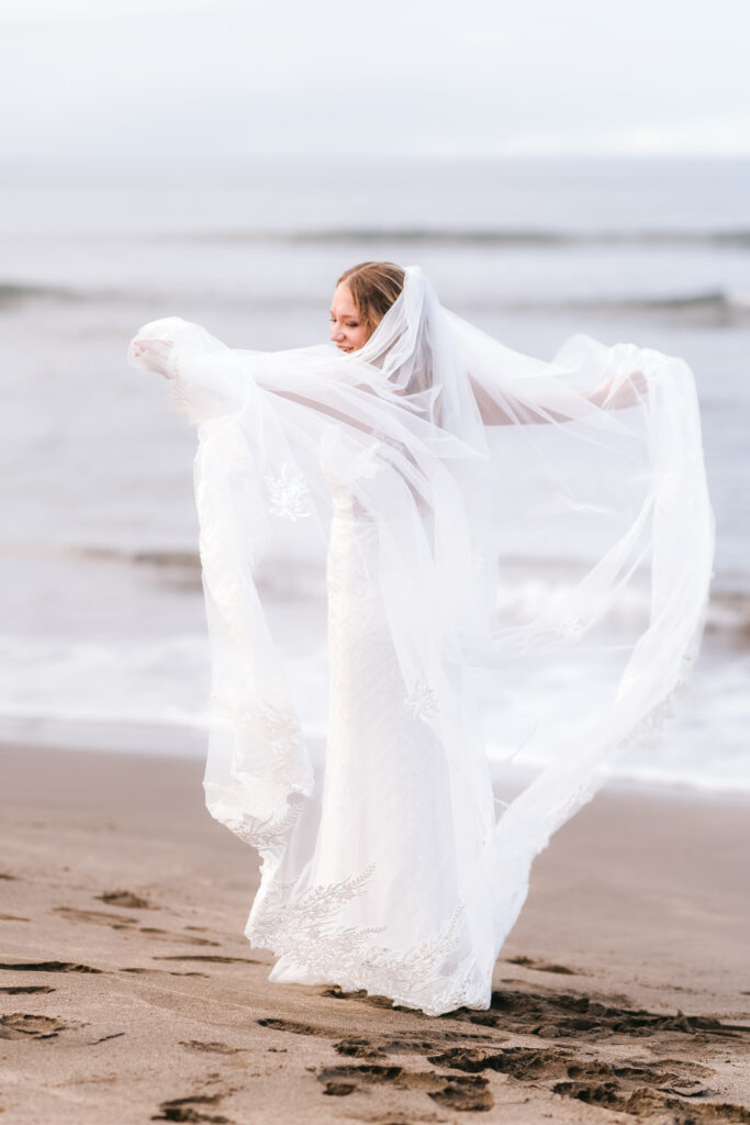 Bride in a fitted lace gown standing on a sandy beach, stretching out her long flowing veil in front of the ocean.

