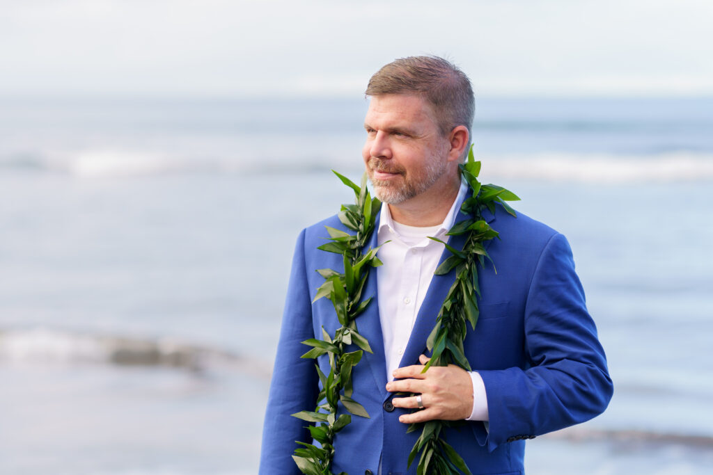 Groom in a blue suit with orange lei standing on a rocky Maui beach at sunset.