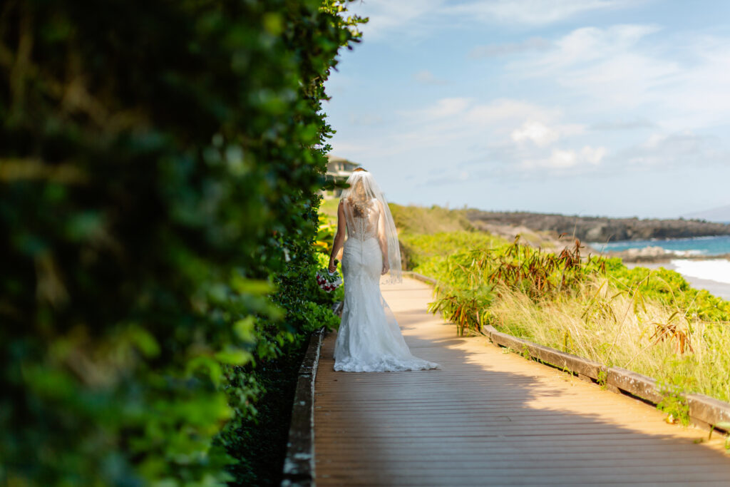 Bride walks alone along a sunlit wooden boardwalk at Ironwoods Beach in Maui during her sunrise elopement, lace train trailing behind her with lush greenery and ocean cliffs ahead.

