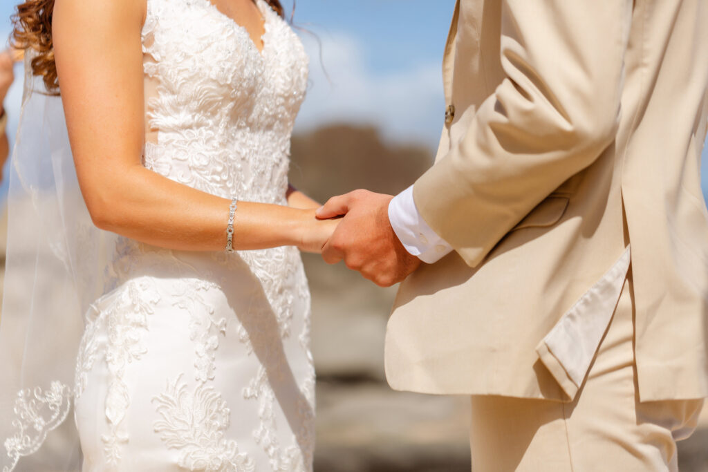 Close-up of bride and groom holding hands during their sunrise elopement at Ironwoods Beach in Maui, her lace gown and his tan suit framed against soft seaside light.