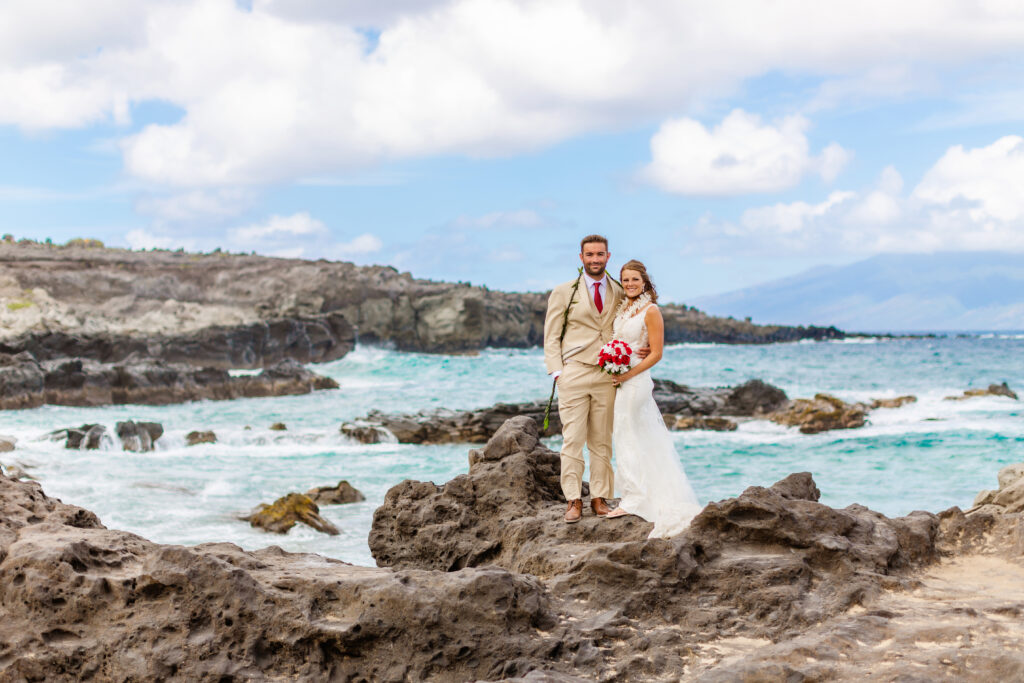 Bride and groom stand together on black lava rocks at Ironwoods Beach in Maui during their sunrise elopement, holding a red and white bouquet with turquoise waves and cliffs behind them.