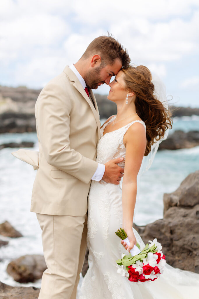 Bride and groom share an intimate moment on the rocky shoreline at Ironwoods Beach in Maui during their sunrise elopement, her lace gown and red-and-white bouquet standing out against the crashing ocean waves. 