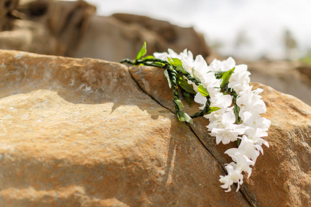 White orchid lei with green leaves resting on a sunlit lava rock at Ironwoods Beach in Maui, captured during a sunrise elopement.