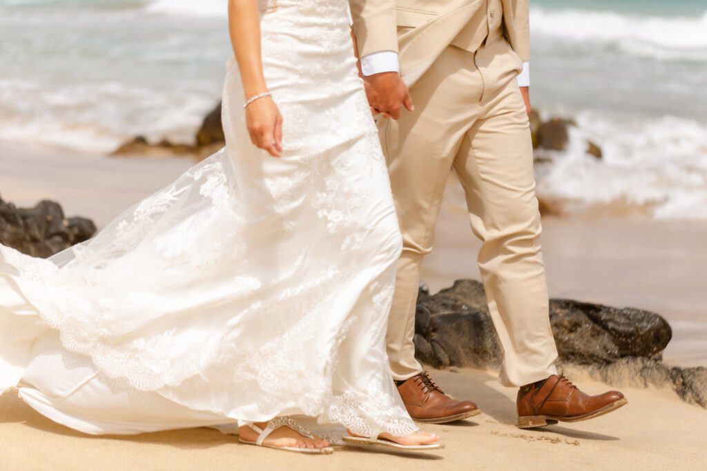 Bride and groom walk hand in hand along the sandy shore at Ironwoods Beach in Maui during their sunrise elopement, her lace gown trailing beside his tan suit and brown dress shoes.
