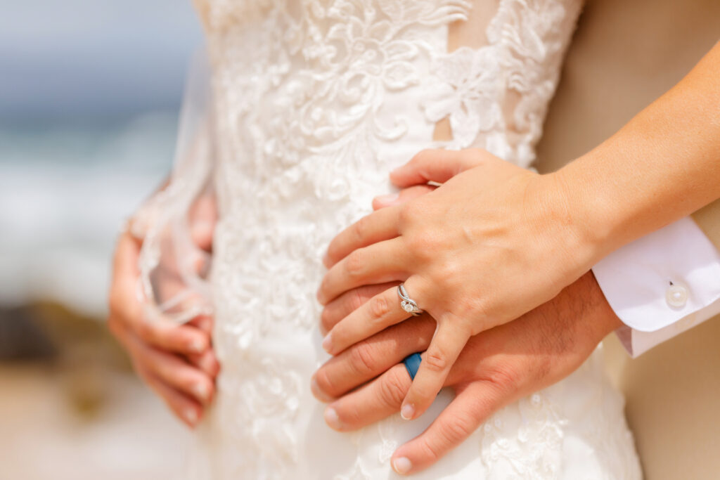 Close-up of the groom’s arms wrapped around the bride’s waist at their sunrise elopement on Ironwoods Beach Maui, highlighting her lace wedding gown and diamond ring against a soft beach backdrop.
