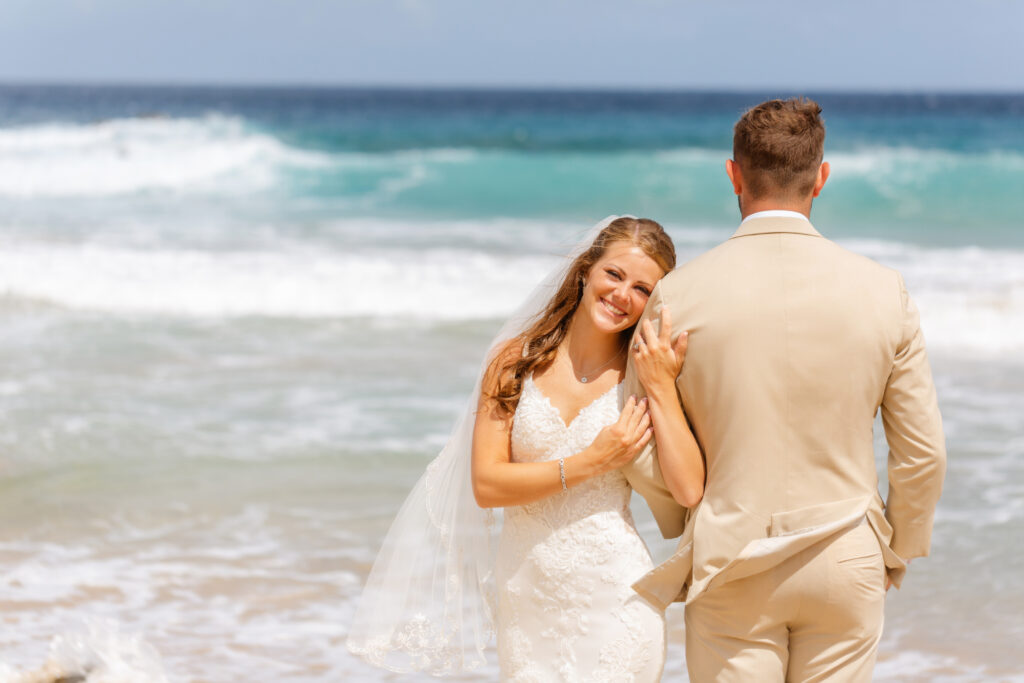 Bride and groom stand arm in arm at the edge of the water during their sunrise elopement at Ironwoods Beach in Maui, facing the waves as her veil and lace gown catch the ocean breeze.