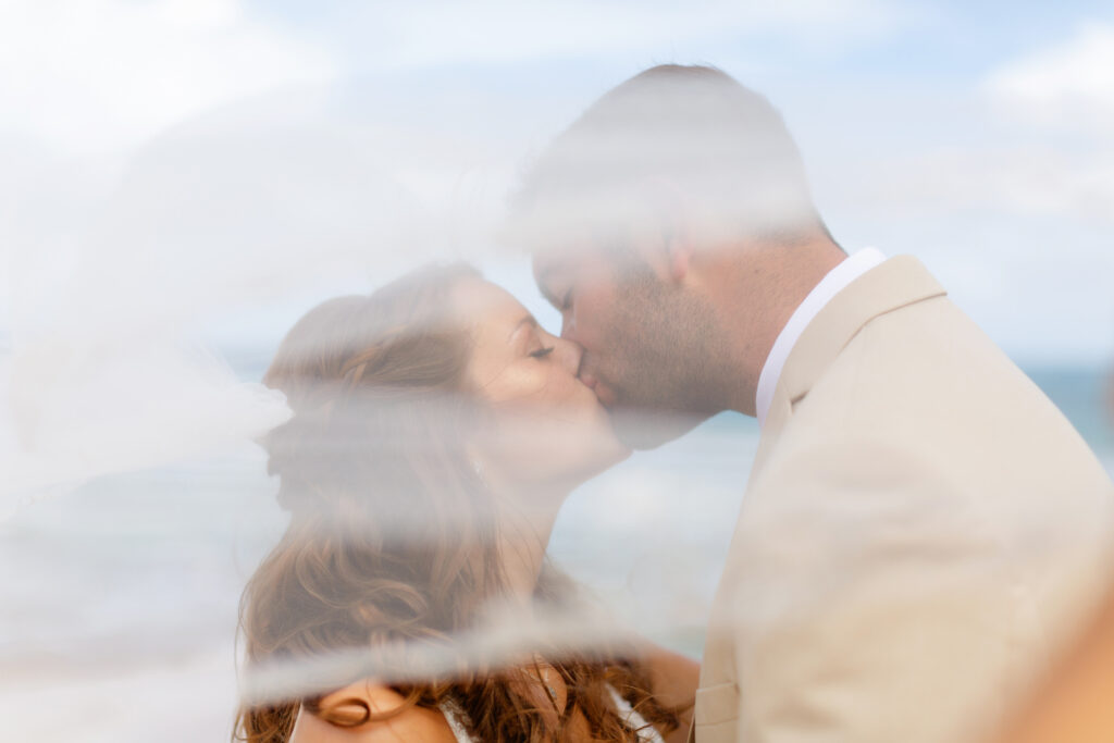 Soft, romantic close-up of the bride and groom sharing a kiss on the beach at their sunrise Ironwoods Beach Maui elopement, framed through the bride’s flowing veil for a dreamy, blurred effect.