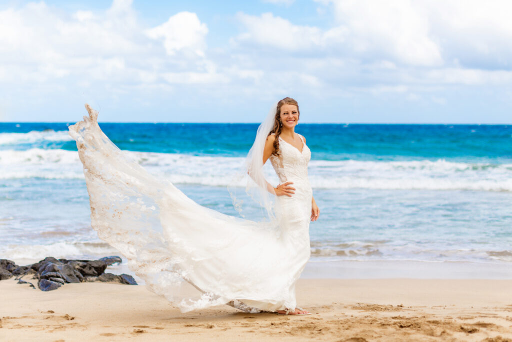 Bride stands on the shore at Ironwoods Beach in Maui during her sunrise elopement, ocean wind catching her lace train and veil as they billow dramatically behind her.