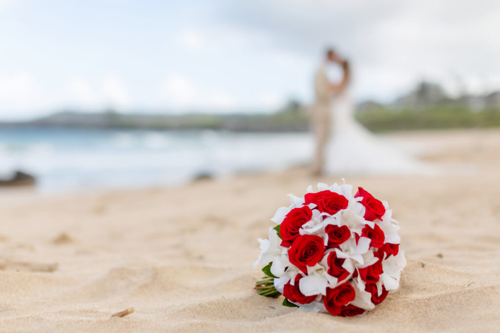 Red and white bridal bouquet resting on the sand at Ironwoods Beach in Maui during a sunrise elopement, with the couple softly blurred in the background sharing a kiss near the shoreline.