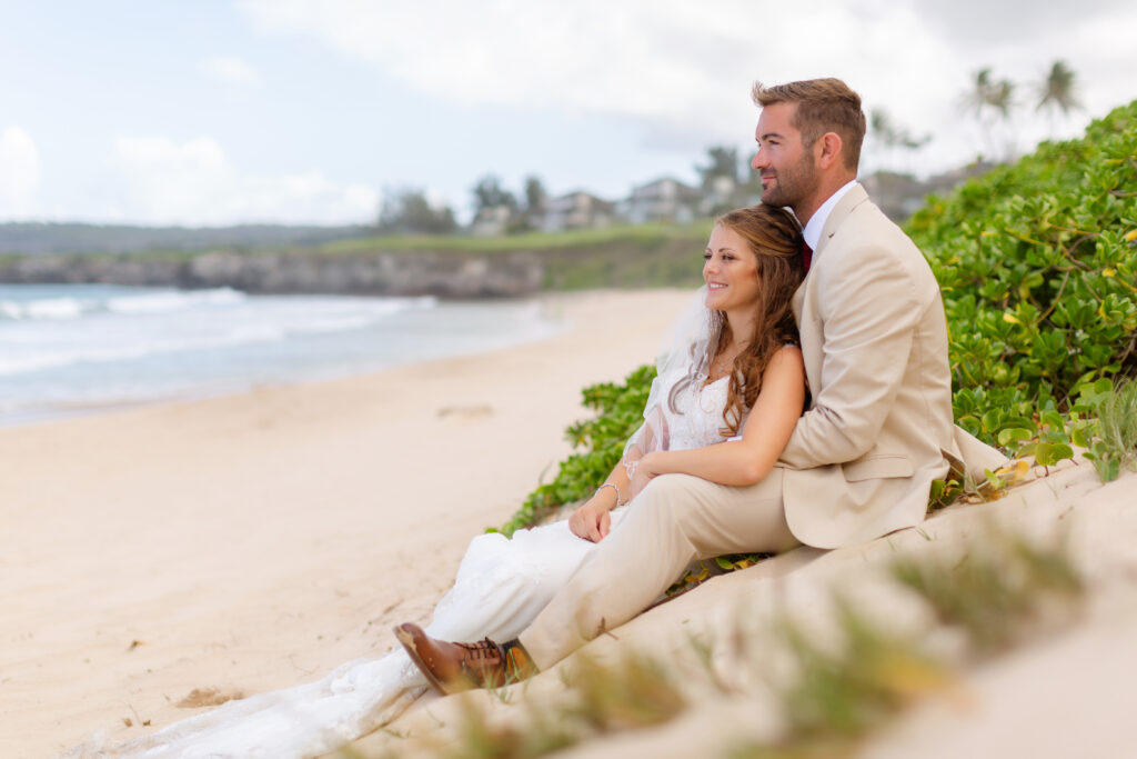 Bride leans back against the groom as they sit together on a sandy dune at Ironwoods Beach in Maui during their sunrise elopement, gazing out at the shoreline and gentle waves.