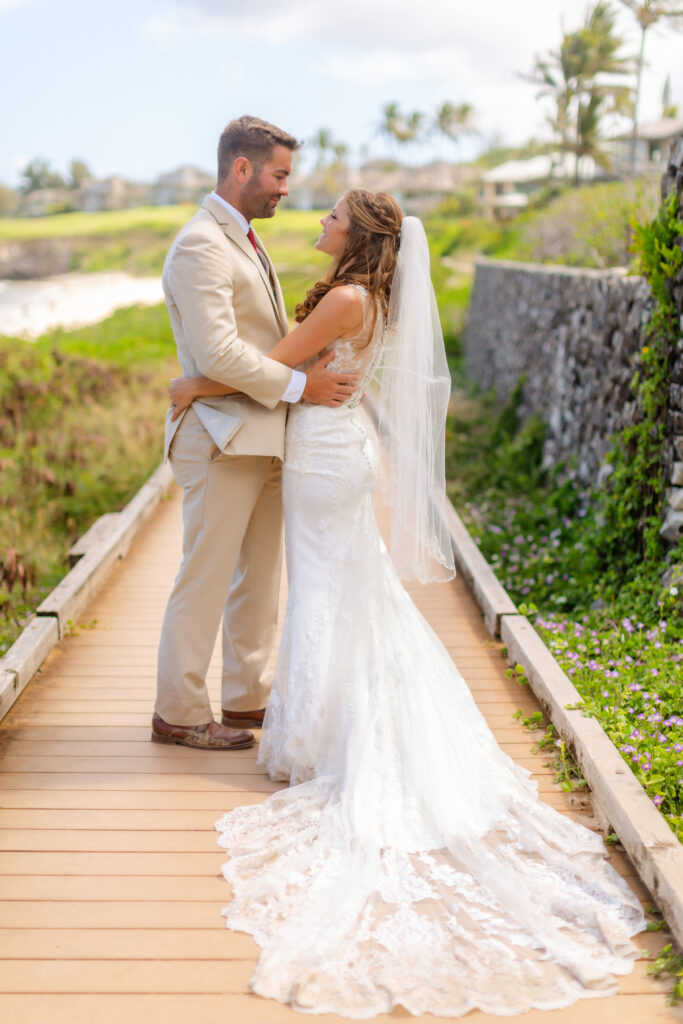 Bride and groom embrace on a wooden boardwalk at Ironwoods Beach in Maui during their sunrise elopement, with her lace train spread behind her, lush greenery and ocean cliffs in the background.