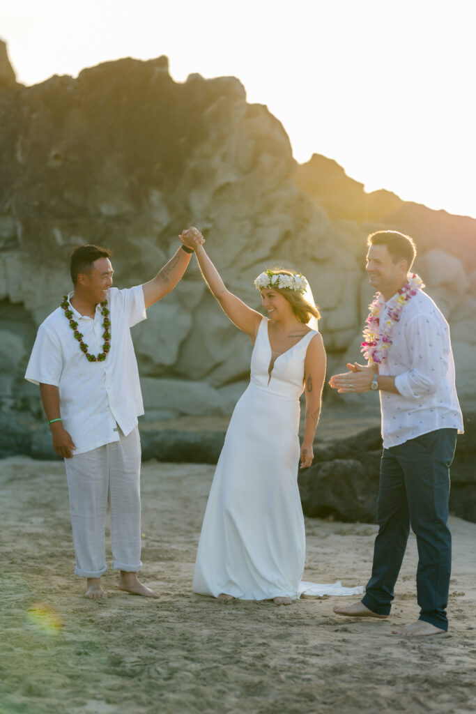 Bride in a simple white gown and flower crown raises her joined hand with the groom, barefoot on the sand at Ironwoods Beach in Maui, while their officiant in a white shirt and lei claps beside them at sunset.