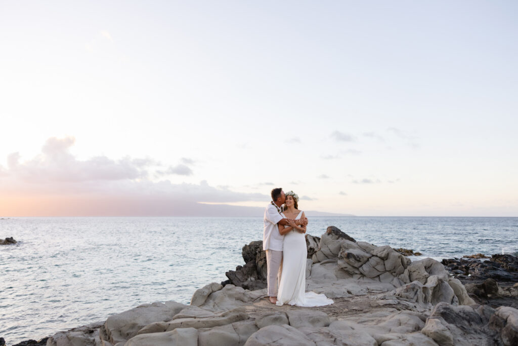 Ironwood Beach Maui wedding ceremony, couple embrace on lava rocks overlooking the ocean at sunset

