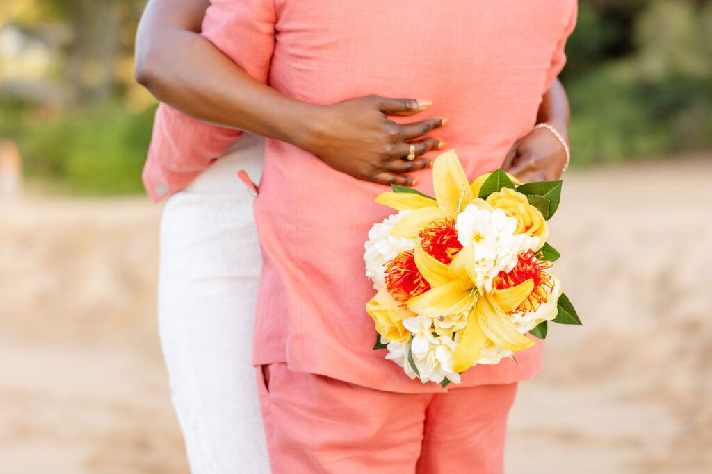 Bride hugging groom from behind while holding a bright tropical bouquet at their Maui beach elopement

