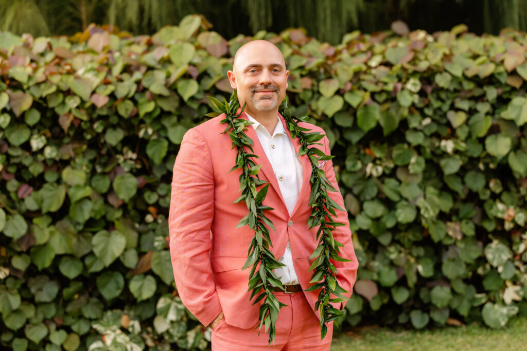 Groom in coral suit wearing a maile lei before his Maui beach elopement