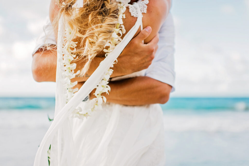 Close-up of a Maui bride and groom embracing on the beach with a floral lei and flowing veil.