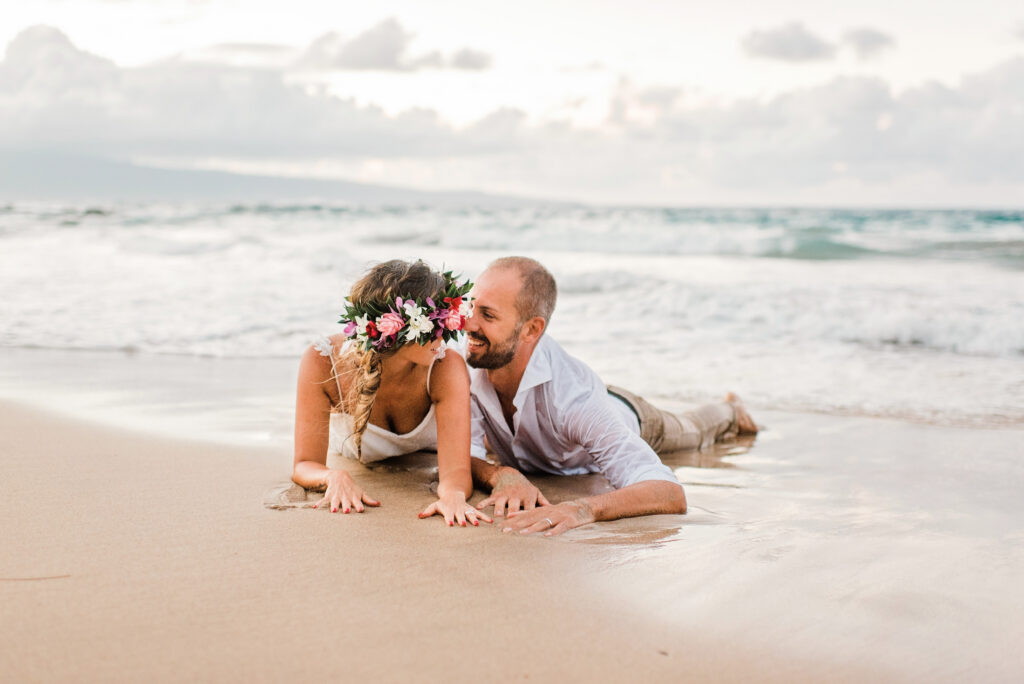 Newlywed couple lying together on the shoreline after their Maui beach wedding with ocean waves in the background.