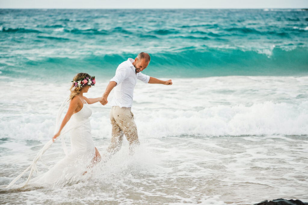 Newlywed couple holding hands and running through the ocean waves after their Maui beach wedding ceremony.