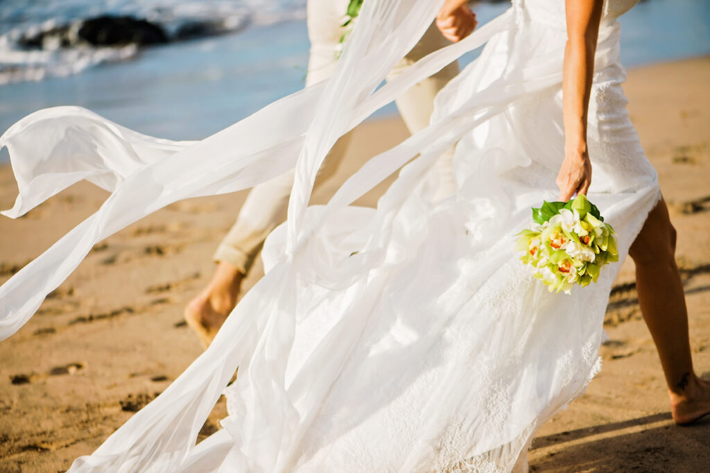 Bride walking along a Maui beach with her wedding dress flowing in the ocean breeze while holding a tropical bouquet.