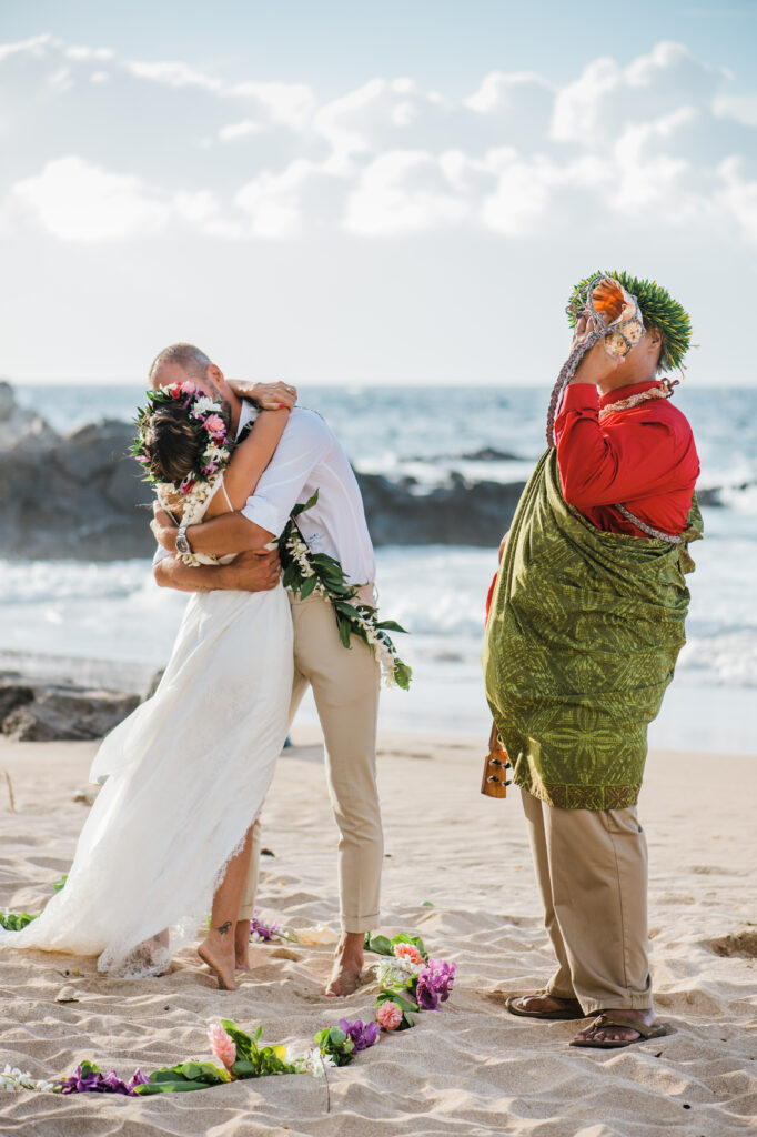 Newly married couple sharing their first kiss during a Maui beach wedding as the officiant blows a conch shell.