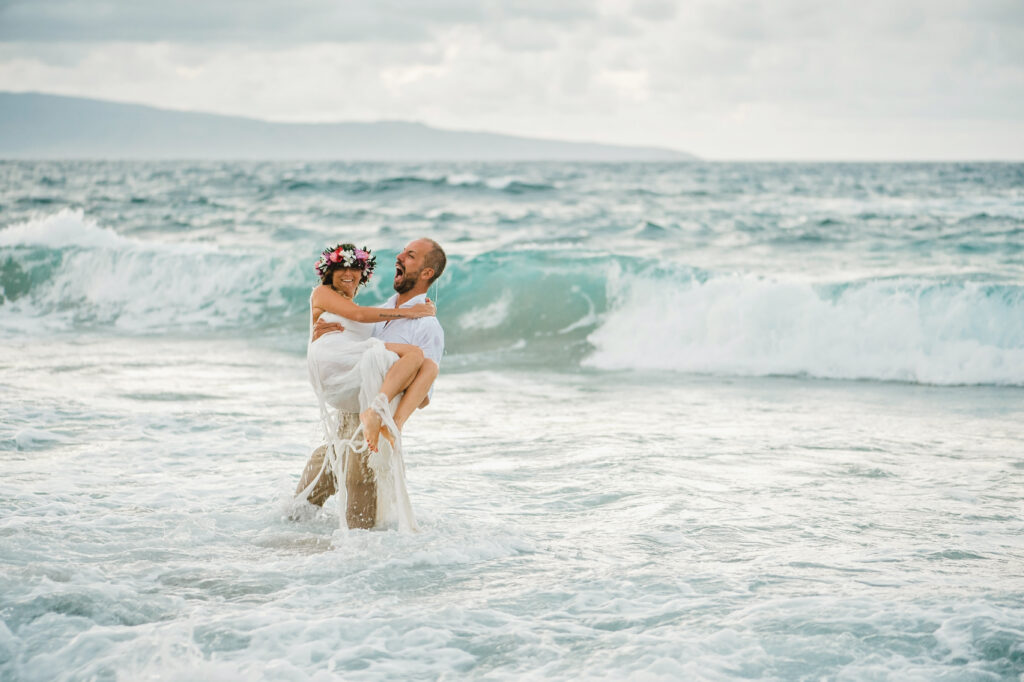 Groom carrying his bride through the ocean after their Maui beach wedding as waves roll in behind them.